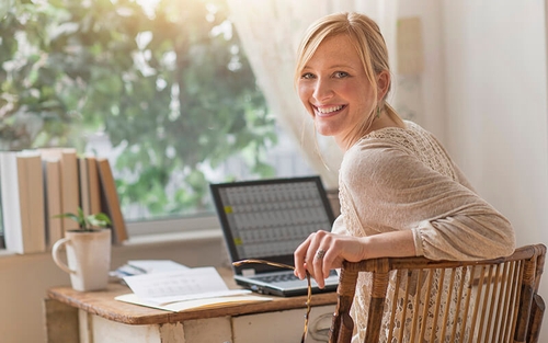 A remote worker smiles while working at an old thrifted desk, showered in natural light by a large window.