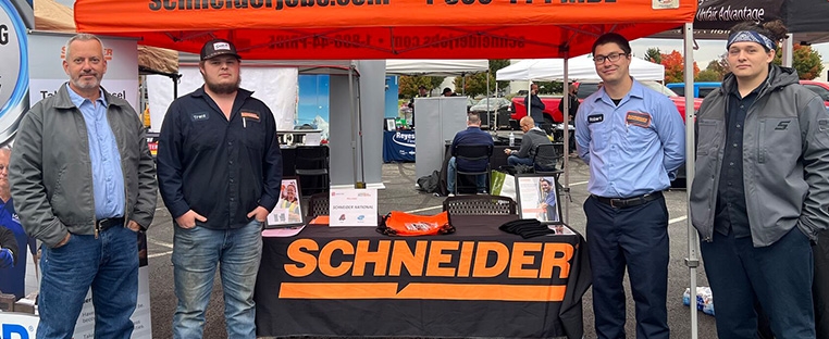 Four Schneider associates, including diesel technicians, stand at a Schneider booth for a hiring event