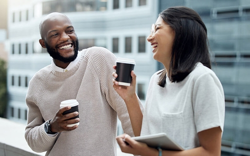 A man and a woman chat while drinking coffee outside an office building.