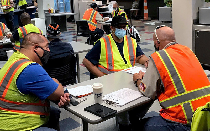 Three men wearing masks and safety vests, sit around a table with papers on it in a cafeteria and have a discussion.