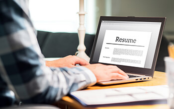 A person wearing a plaid shirt sits at a table and works on their resume on their laptop.