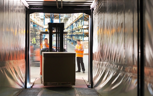 In a warehouse setting, while one warehouse associate takes a pallet off a semi-trailer using a forklift, another warehouse associate looks at a tablet.