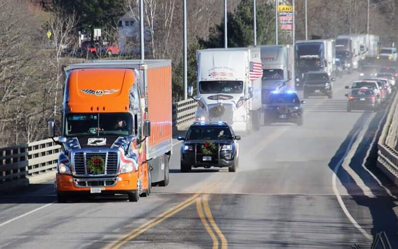 Jon leads the police escorted Wreaths Across America convoy across a bridge in his specially wrapped Ride of Pride truck. He is followed by a line of police cars and other semi-trucks hauling trailers full of wreaths.
