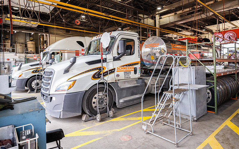 Two Schneider company trucks hauling tanker and van trailers sit in the service bays of a Schneider shop awaiting service.