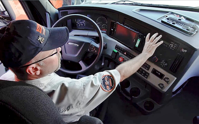 Driving Instructor Brett waves his hand in front of the dashboard of a 2021 Freightliner Cascadia semi-truck.