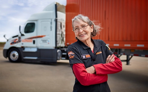 A mature Schneider truck driver poses in front of her company truck, which is hauling an orange container.