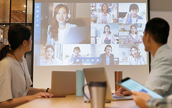 Two individuals in an office setting are having a meeting with a group of associates who are virtual on a large screen.