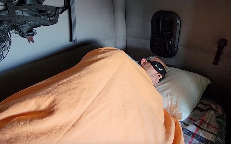 A truck driver sleeping under an orange blanket with a sleep mask, in the sleeper berth of their semi-truck.