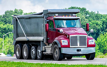 A red and silver dump truck drives on a road surrounded by green trees.