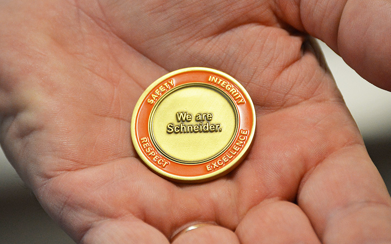 The backside of the Schneider Value Coin features the words "We are Schneider." inside an orange ring that lists Schneider's core values of Safety, Integrity, Respect and Excellence.