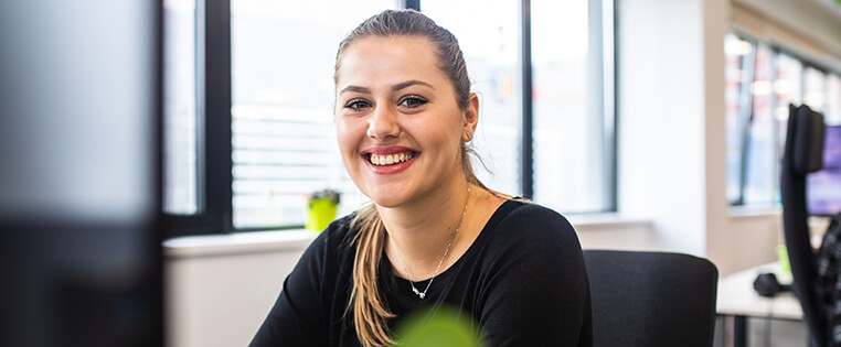 An MLS office associate sitting at her desk, smiling at the camera, with a large window behind her.