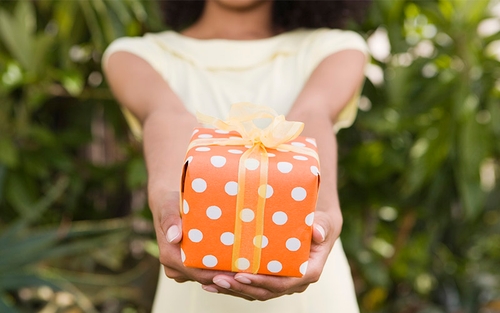 A woman holding out and a gift wrapped in orange wrapping paper and a yellow bow. 