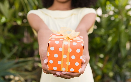 A woman holding out and a gift wrapped in orange wrapping paper and a yellow bow.