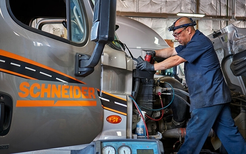 A Schneider diesel technician works on the front of a grey Schneider trailer in a shop.