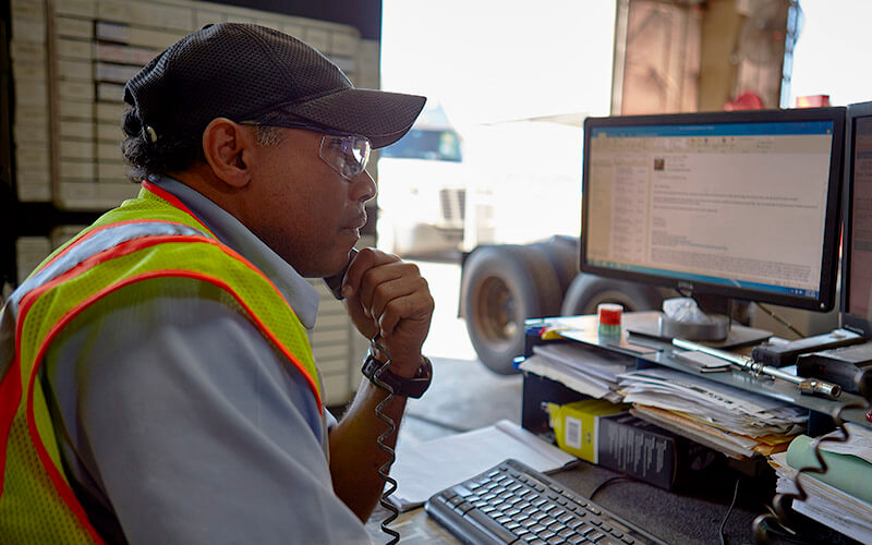 A diesel shop manager holds a phone to his ear while looking over an email at a desk to the side of a service bay, where there is a truck parked and awaiting service.