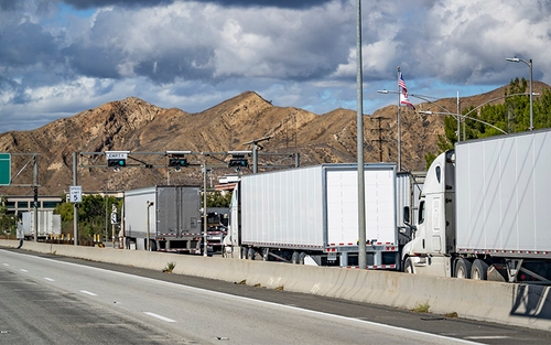 Several white semi-trucks waiting in line at a weigh station. In the background, there are large brown mountains under a partly cloudy sky with an American flag flying atop a pole