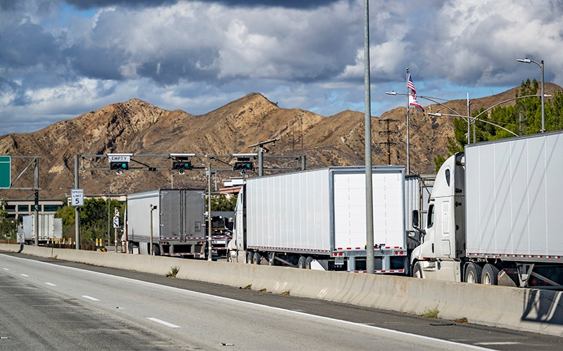 Several white semi-trucks waiting in line at a weigh station. In the background, there are large brown mountains under a partly cloudy sky with an American flag flying atop a pole 