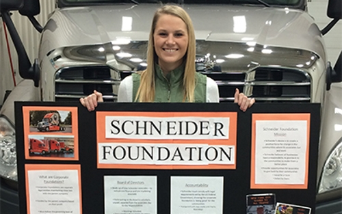 Schneider intern Rachel Steffel holds up her research project display on the Schneider Foundation while standing in front of a semi-truck.
