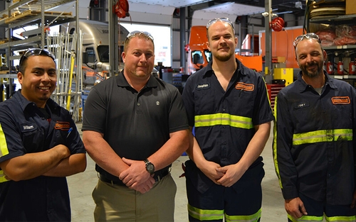 Four men stand who are smiling stand in a line within a Schneider shop. Three of the four are wearing diesel technician outfits while the third wears khaki pants and a grey polo shirt.