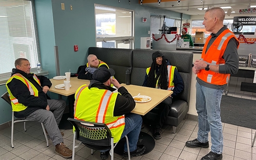 A Schneider driver instructor talking to a group of new drivers in a facility cafeteria.