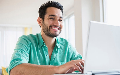 A male associate in a turquoise button-down shirt is working from home and smiles cheerfully while typing on his laptop in a bright, sunlit room.
