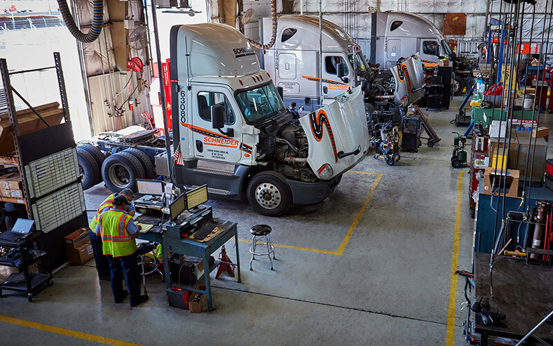 Two diesel technicians review paperwork in a Schneider diesel technician shop with three company trucks parked in service bays with hoods open.