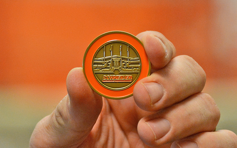 A front view of the Schneider Value Coin shows the company logo with an etching of the corporate headquarters.