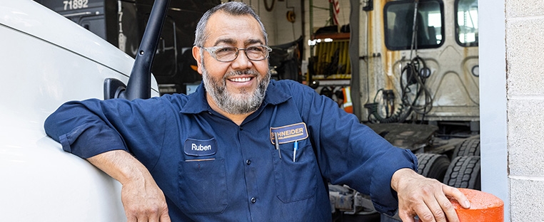 A Schneider diesel technician leaning against the hood of a semi truck in front of a maintenance bay