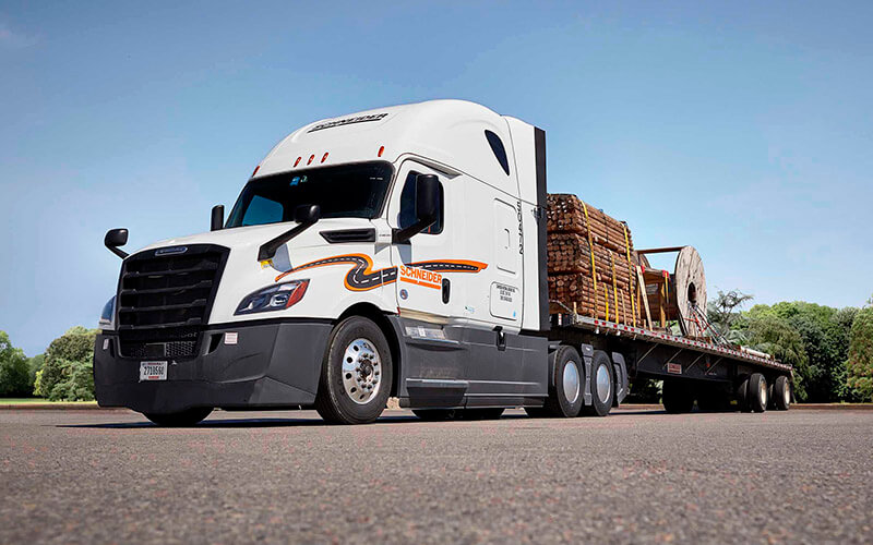 A white Schneider semi-truck hauling a flatbed trailer holding wood is parked in a lot.