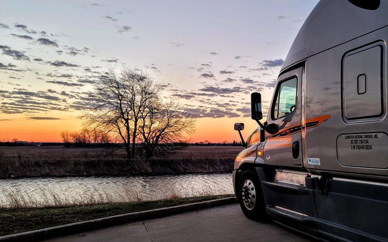 A grey Schneider semi-truck is parked in a lot that overlooks a body of water and a colorful sunset.