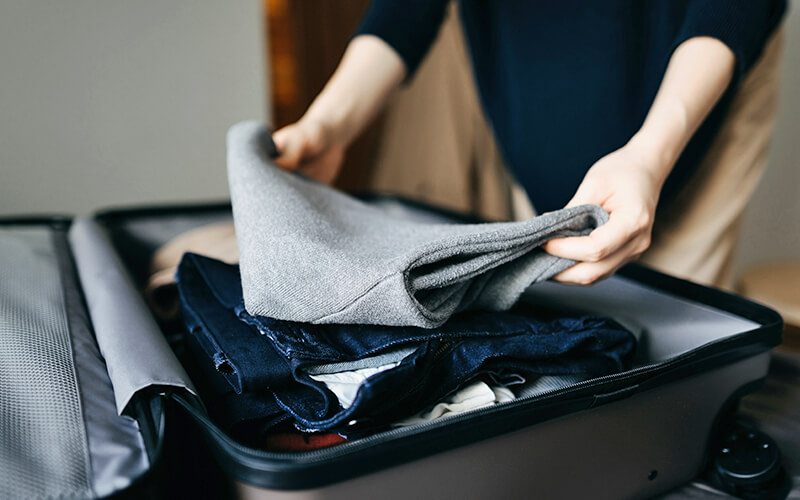 A man wearing khaki pants and a black long sleeve shirt packs a grey suitcase with various clothes, including a grey sweater and dark blue jeans.