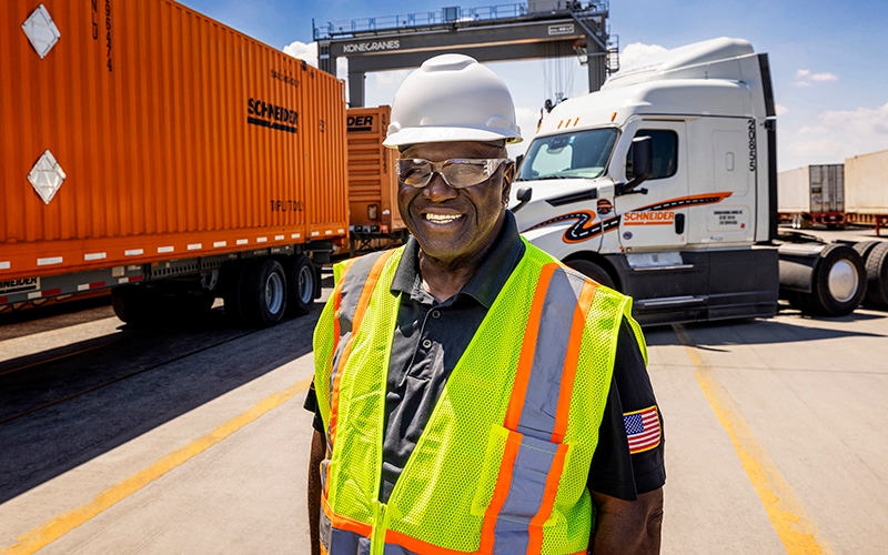 An intermodal driver standing in a railyard wearing a high-visibility safety vest, with semi-trucks and orange shipping containers in the background.