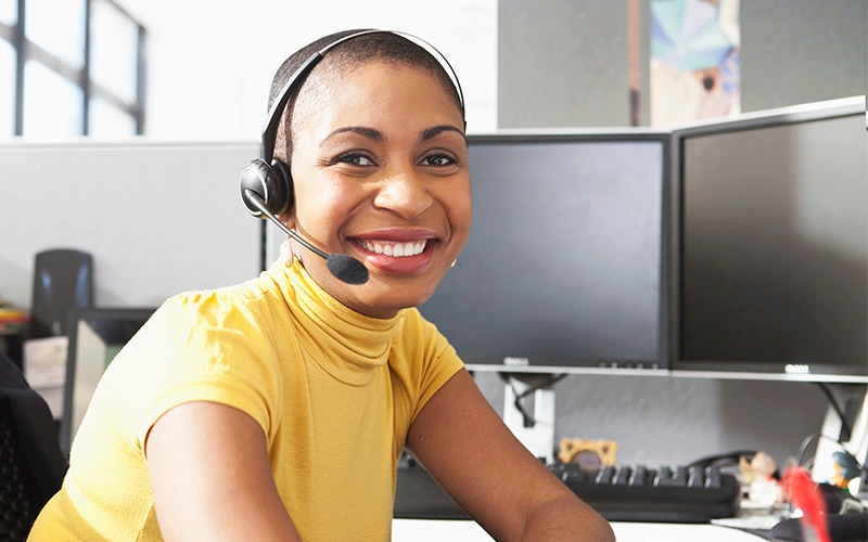 A woman in a yellow shirt and headset working at a desk with multiple monitors. 