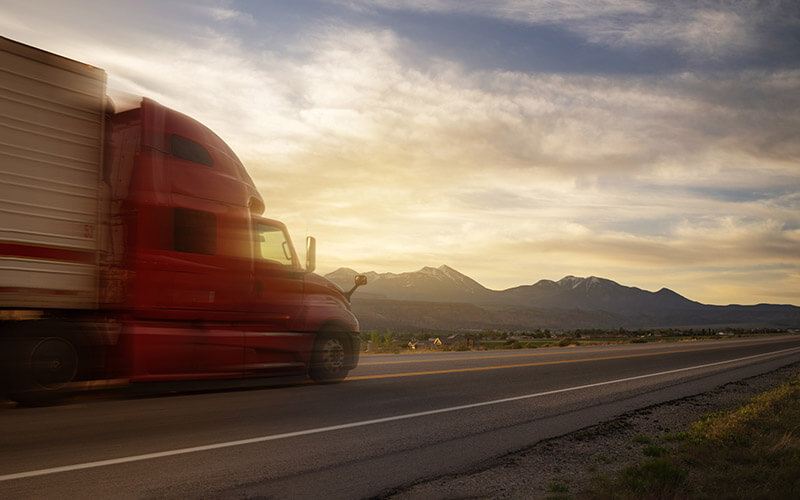 A red semi-truck hauling a trailer appears to be traveling quickly on a highway overlooking a mountain range.