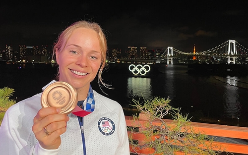 At night time, Krysta Palmer stands in front of a ridge in Tokyo and holds her bronze Olympic medal.