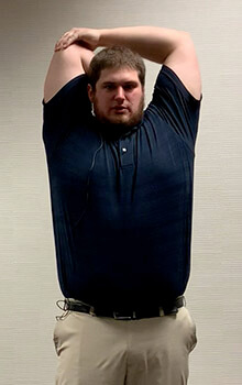 A physical therapist demonstrates stretching his elbow by bringing his arms overhead, grasping his elbow with the opposite arm and bending his elbow to reach down his back.