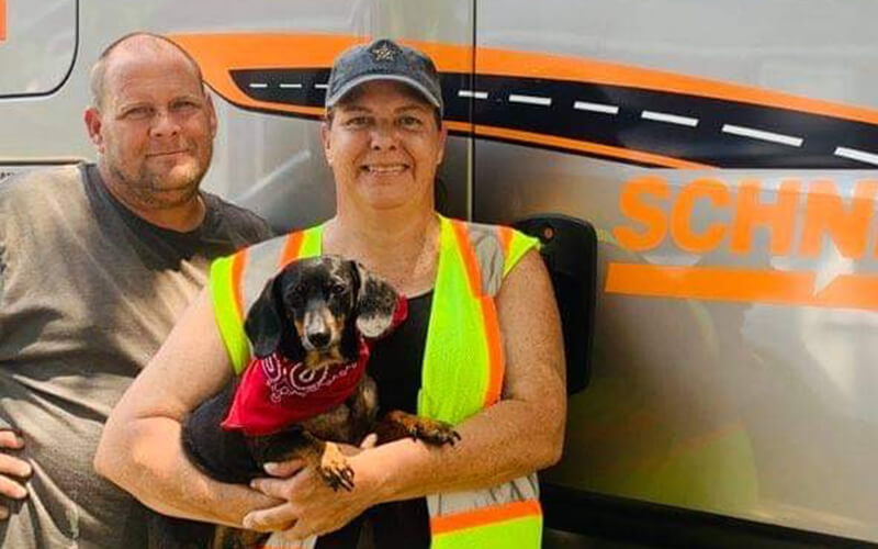 Calvin and Rhonda smile in front of their Schneider semi-truck. Calvin stands behind Rhonda, who is holding their dachshund, Harley. Calvin is wearing a brown shirt, Rhonda is wearing a cap and neon vest and Harley has a red bandana.