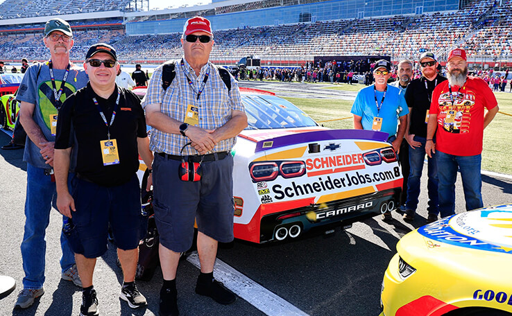 A group of Schneider drivers near the race track at a NASCAR event, standing near the backside of a race car that has the Schneider logo and "schneiderjobs.com" written underneath it with the seating area behind them