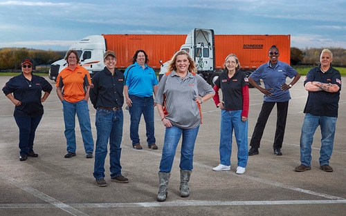 A diverse group of female Schneider truck drivers standing in a parking lot in front of two intermodal company semi-trucks.