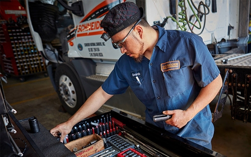 A Schneider diesel technician looking through his toolbox in a shop setting with a Schneider tractor parked in the background.