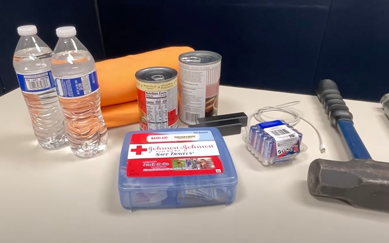 Truck driver essentials laid out on a table: bottled water, a rolled-up orange blanket, canned food, a first aid kit labeled ‘Johnson & Johnson Safe Travels,’ batteries and a flashlight.
