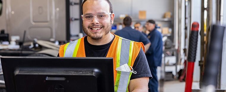 A Schneider diesel technician stands at a computer inside a maintenance shop, with other technicians working in the background