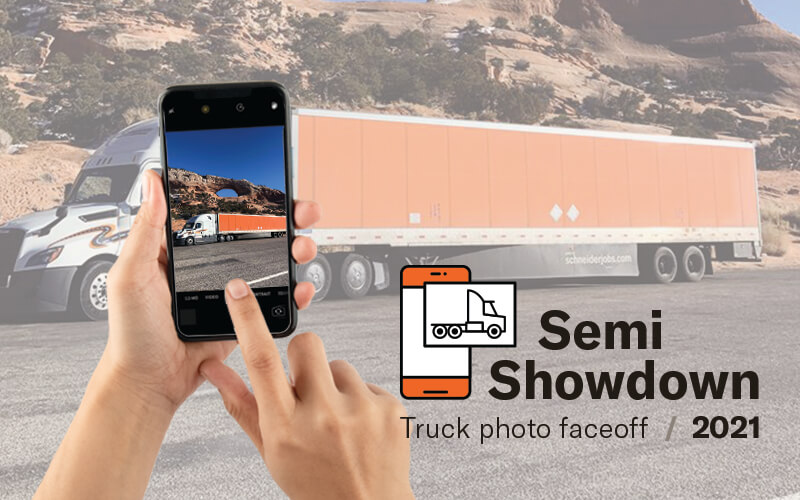 A hand holding a phone takes a picture of a Schneider semi-truck. The bottom right corner says "Semi Showdown truck photo face off 2021."
