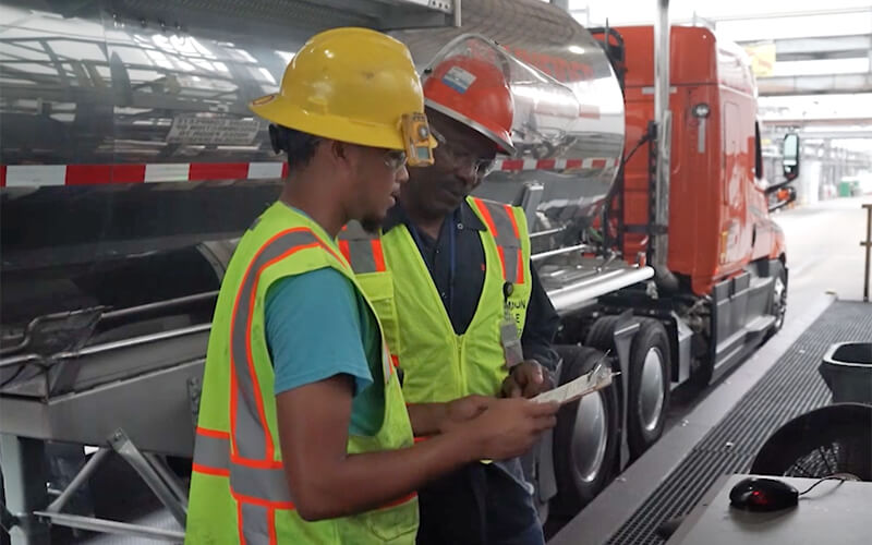 A Schneider tanker driver in a yellow hard hat and green safety vest talks with a Schneider diesel technician who is wearing a red hard hat and green safety vest.
