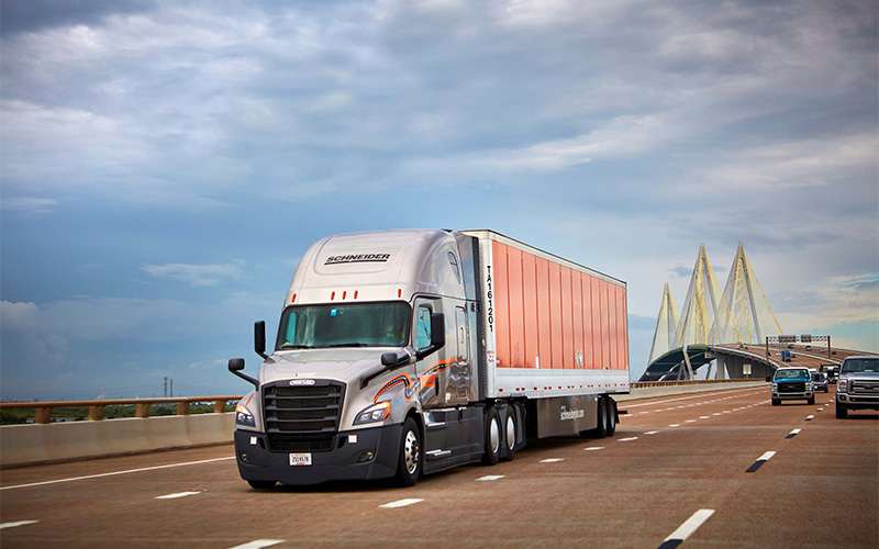 A grey Schneider semi-truck hauling an orange trailer crosses a large highway bridge.