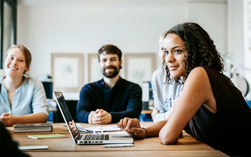 A group of people seated around a table in a meeting or collaborative work environment. Laptops and documents are visible on the table. The room has bright natural lighting.