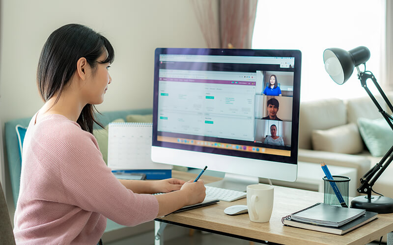 A young woman engages with her coworkers during a virtual meeting on the computer while she works from home.