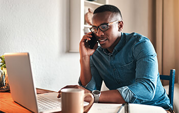 A man in a jean shirt sits at a table and talks on a cell phone with a laptop open in front of him.