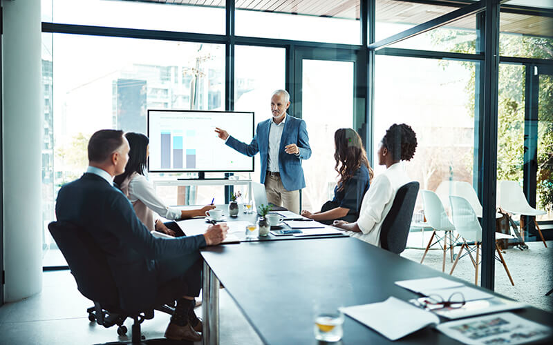 An associate gestures at a graph on a large screen at the end of a conference table lined with colleagues 