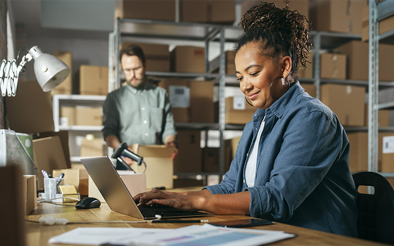 A woman typing on a computer and a man wrapping a package in the background.